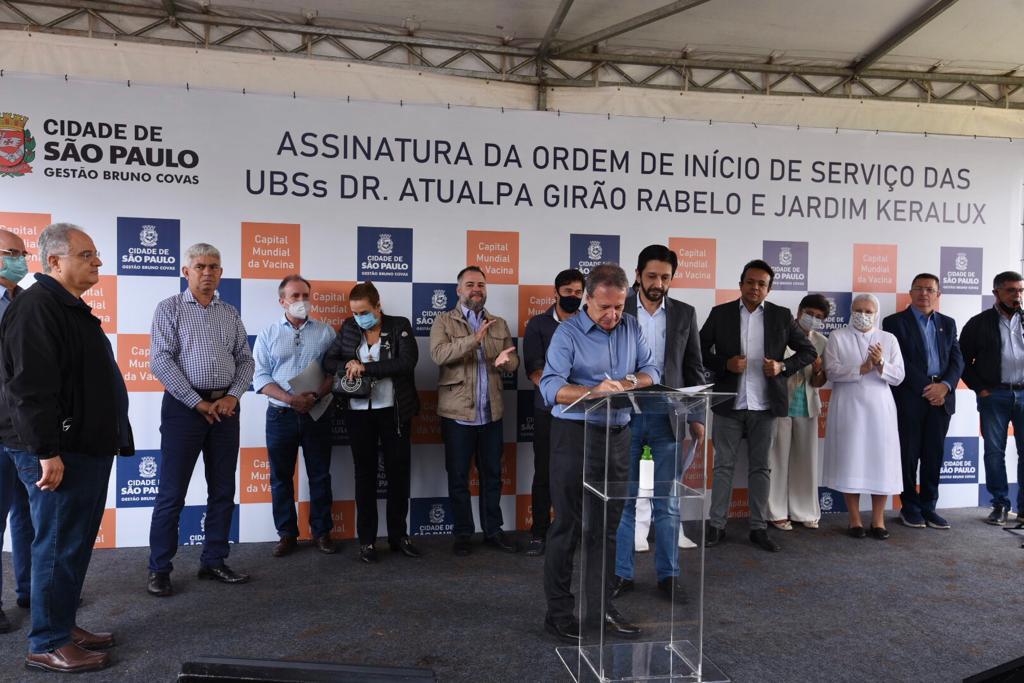Na imagem há diversas pessoas em frente a um painel da prefeitura de São Paulo. À frente das pessoas está o secretário Edson Aparecido e o prefeito Ricardo Nunes.
